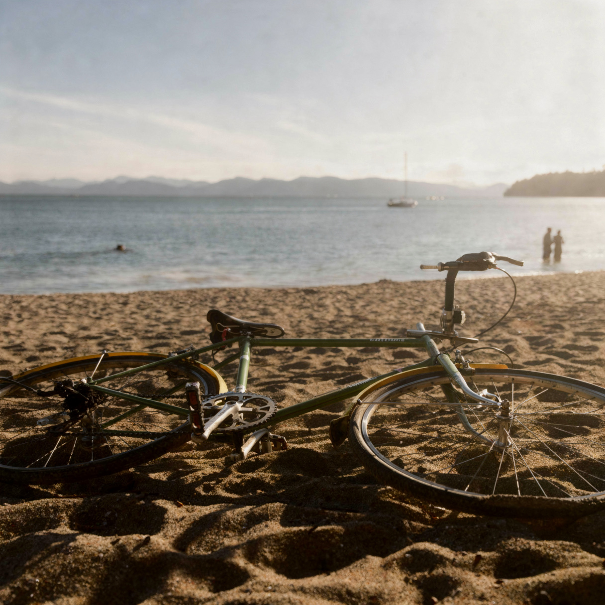A bicycle lying on a sandy beach at the water's edge, calm sea and distant mountains in soft morning light, two people wading in the water