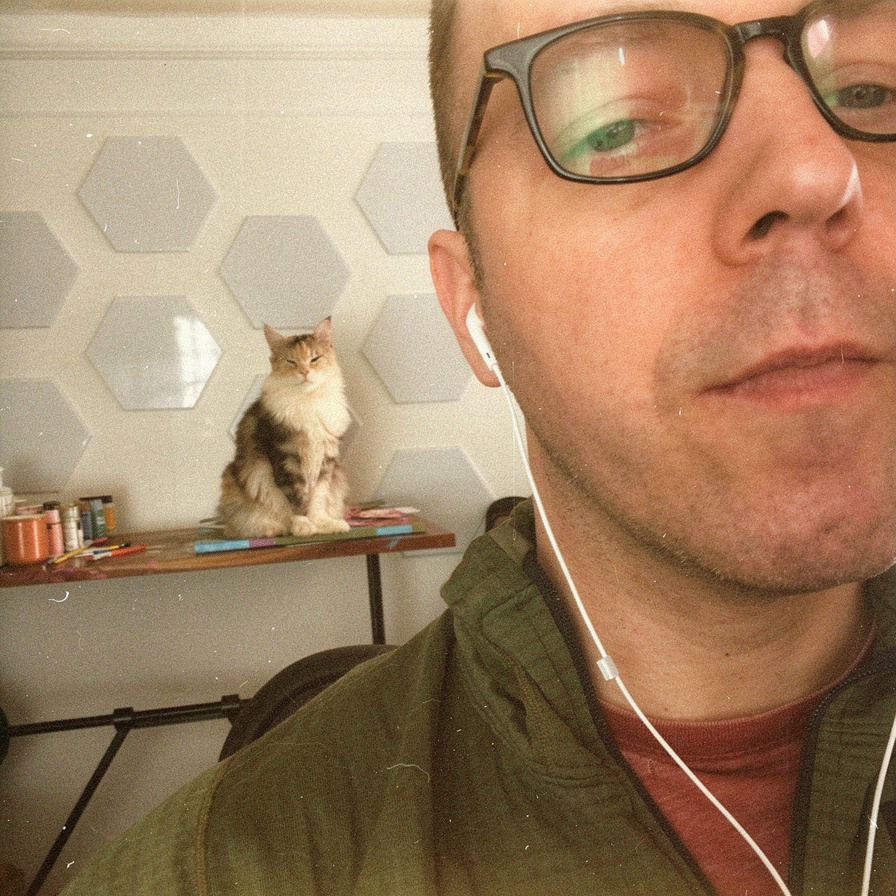 A close-up selfie wearing glasses and earbuds, with a cat sitting on a desk in the background